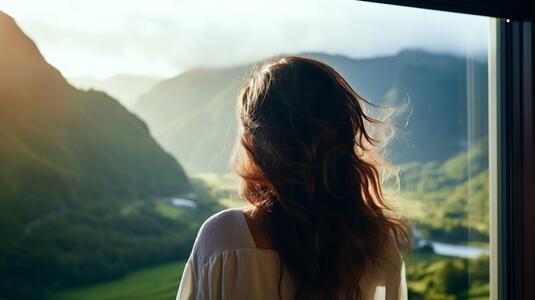 woman with wavy brown hair stands by a large window, gazing out at a breathtaking mountain landscape bathed in golden sunlight. She wears a white off-the-shoulder top, and her hair is gently tousled by the breeze. The scene exudes a sense of peace, reflection, and connection with nature.